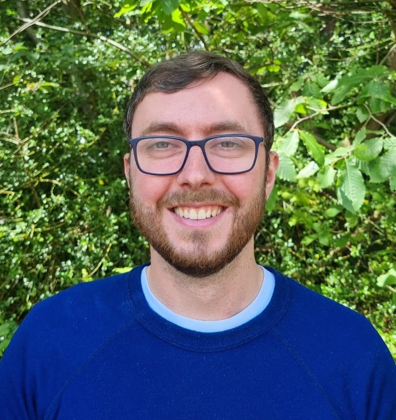 Cam A man with glasses and a blue shirt smiles outdoors with green foliage in the background.