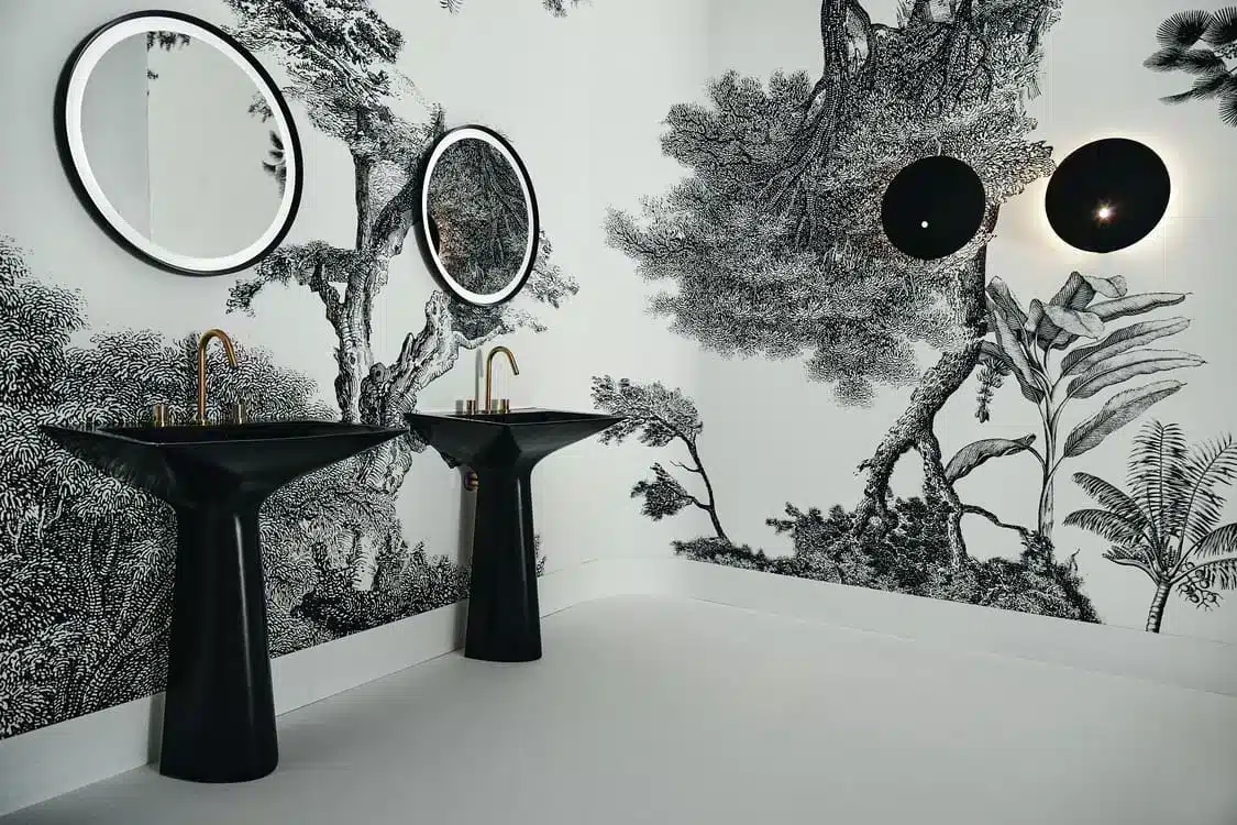 IdealStandard-Journey Two black pedestal sinks with round mirrors and a black-and-white forest wallpaper.