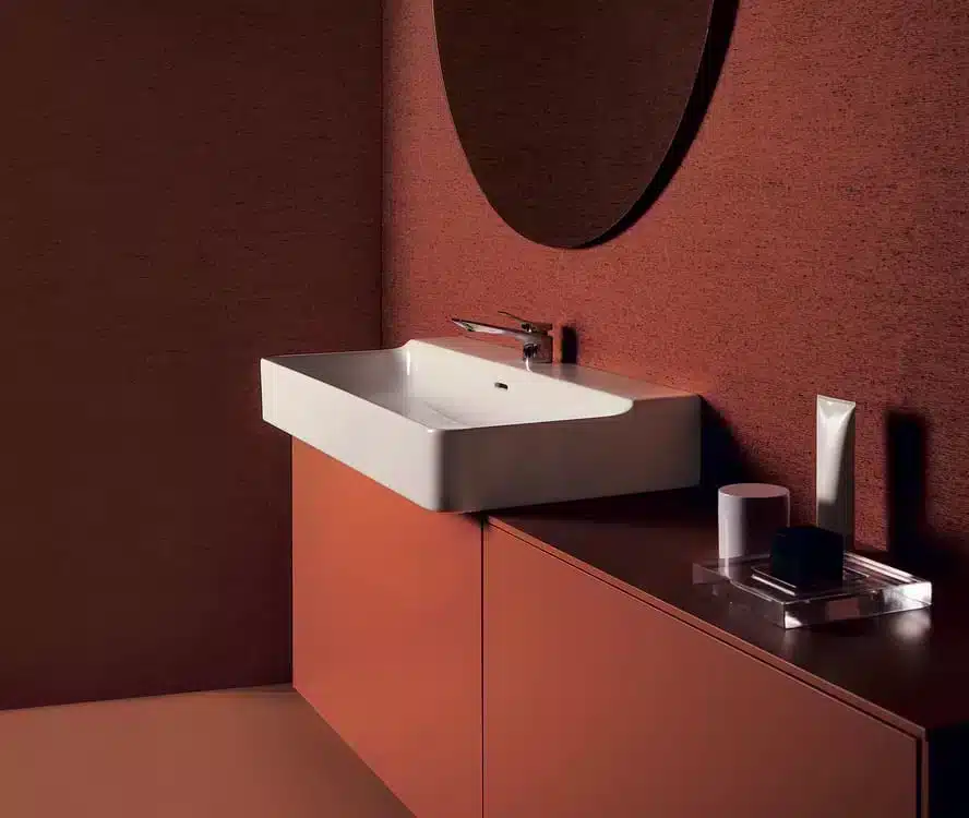 IdealStandard-TheChallenge Modern bathroom with a white rectangular sink, round mirror, and brown cabinets against brown walls.