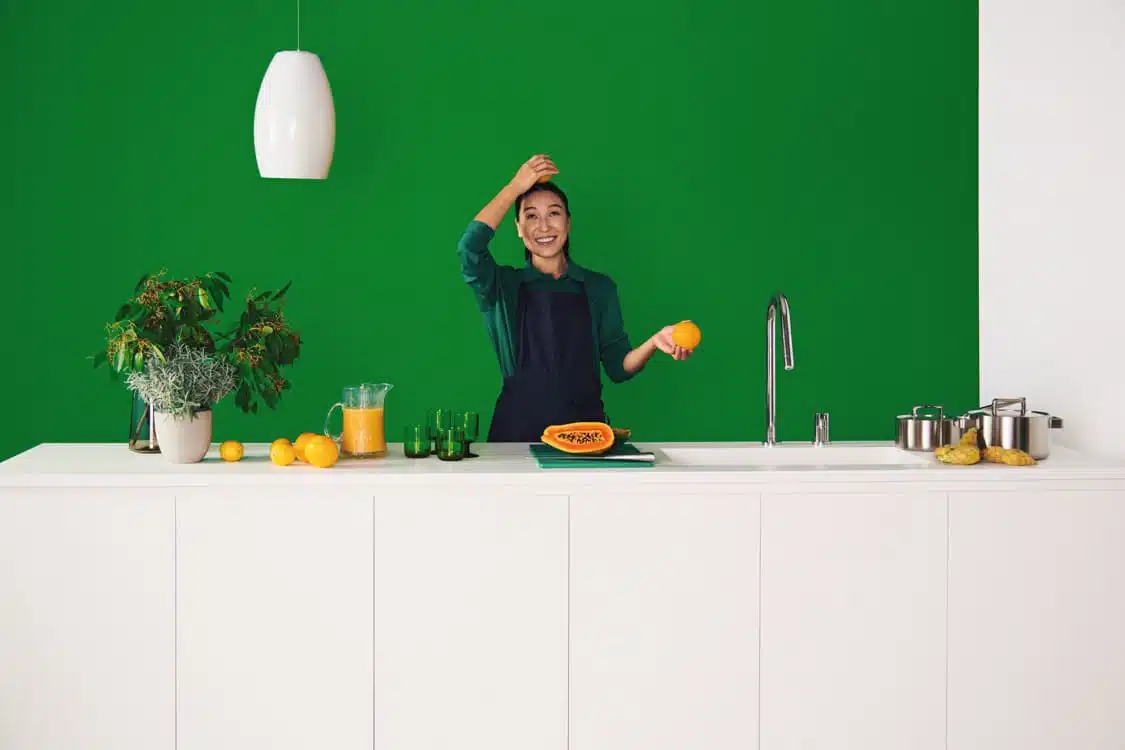 IdealStandard-TheSolution A woman in a kitchen with a green wall, holding an orange and smiling by a white counter and sink.