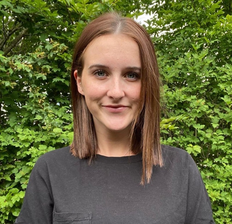 Leah Woman with straight brown hair and a black shirt stands outdoors in front of green bushes.