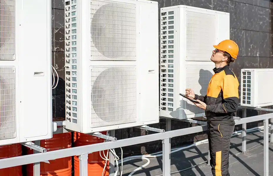Technician inspecting outdoor heat pump units on a building wall, wearing an orange hard hat and uniform.