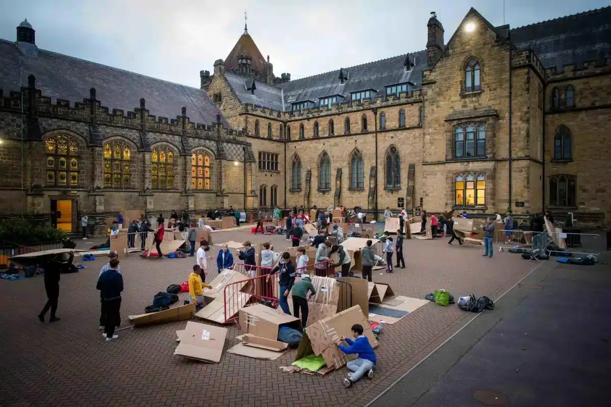 Students build cardboard shelters in a school courtyard outside a large historic stone building at dusk.