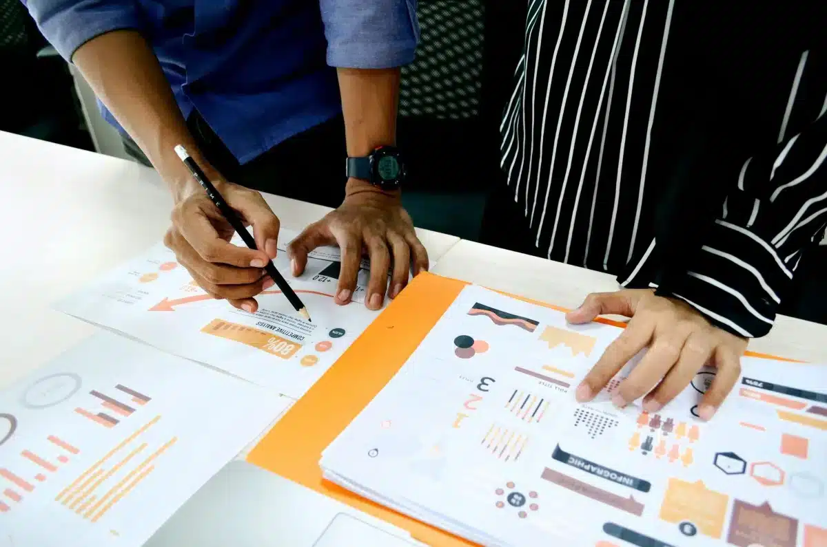 Two people reviewing and discussing colorful charts and graphs on printed sheets at a desk.