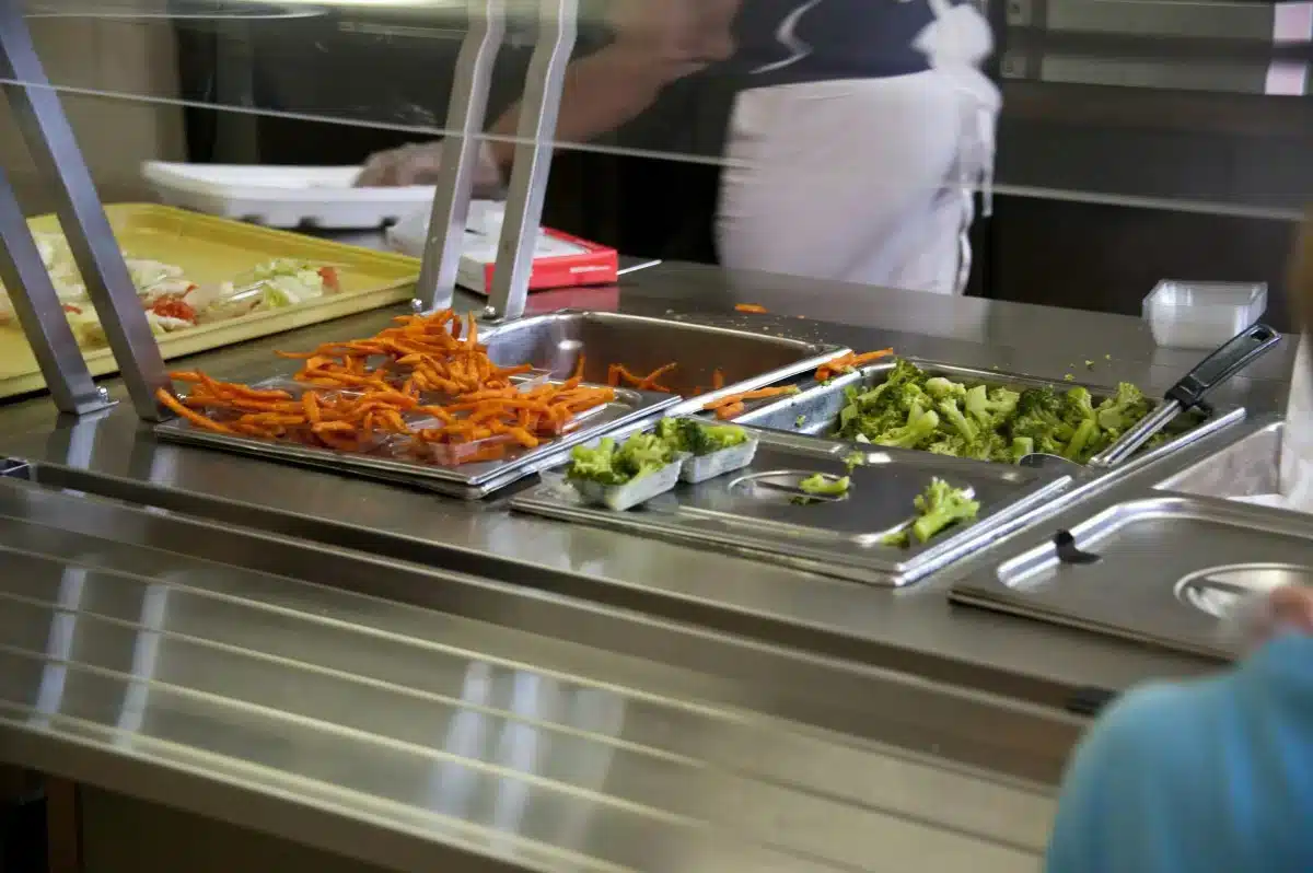 A cafeteria food counter with trays of carrots and broccoli behind a glass shield.