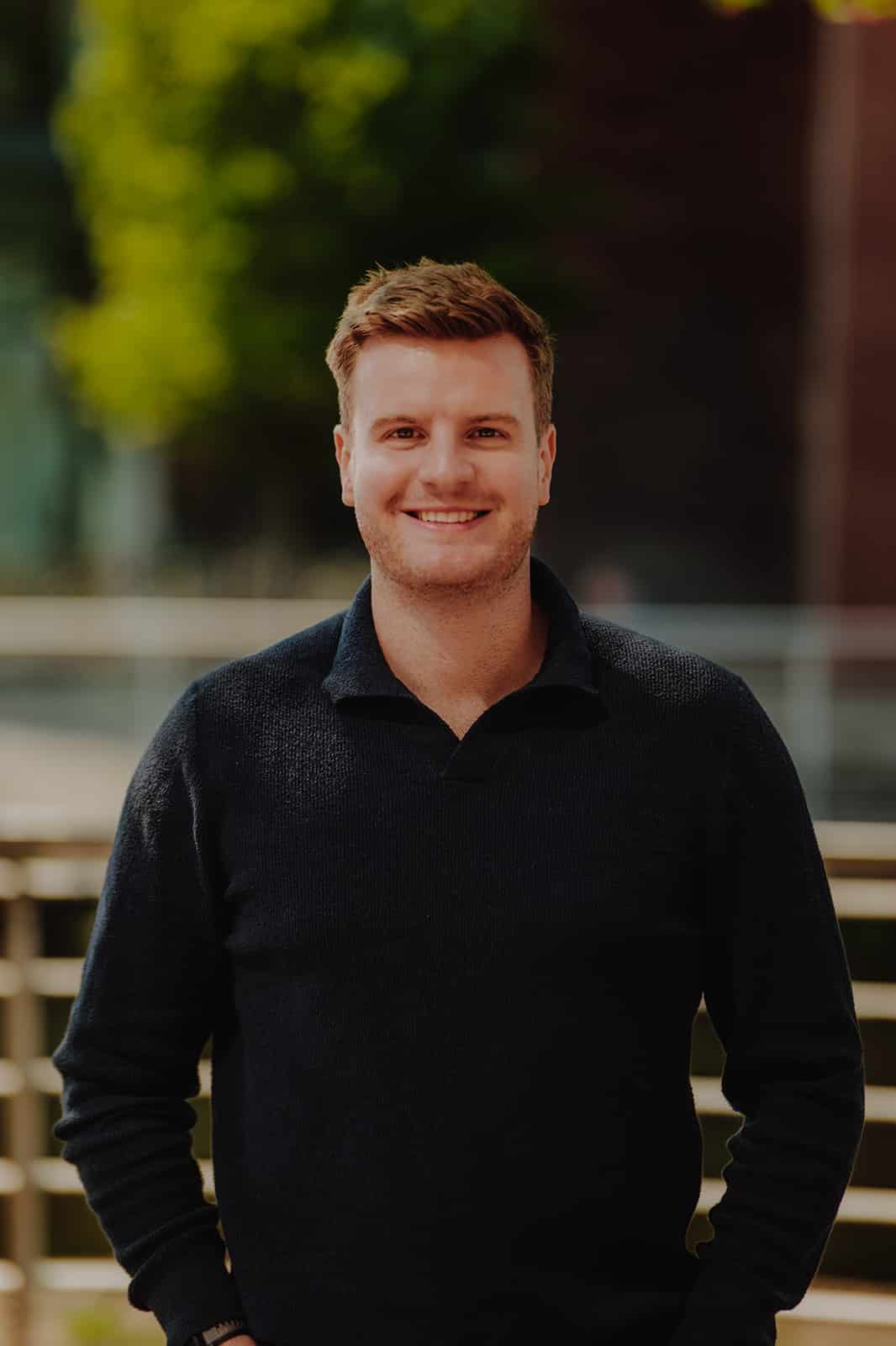 MY CARBON Smiling man in a dark sweater standing outdoors with trees in the background.