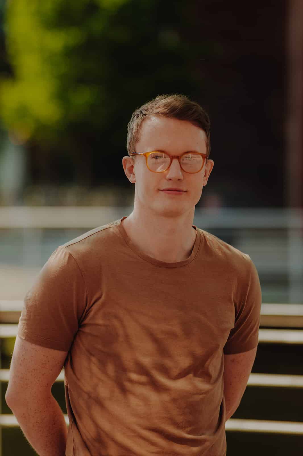MY CARBON A man with glasses and a brown shirt stands outdoors with blurred greenery in the background.