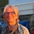 Smiling woman with glasses and short hair outdoors, wearing a scarf and denim jacket.
