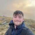 A person smiling outdoors on a grassy, rocky hillside at sunset.