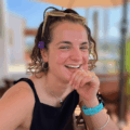 A smiling woman with sunglasses on her head and a flower in her hair sits at an outdoor café.